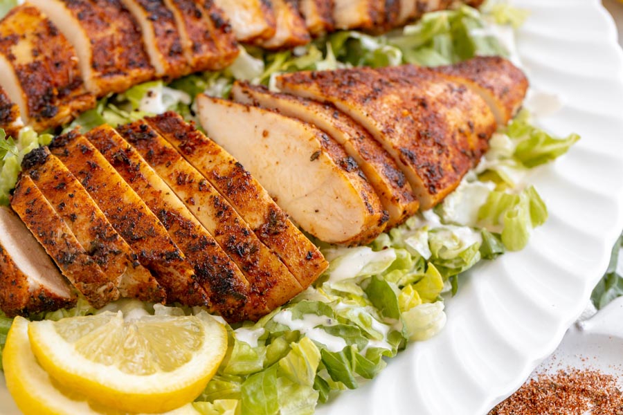 Sliced blackened chicken breast over Caesar salad on a white platter with sliced lemon nearby.