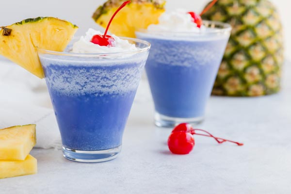 two frozen blue cocktails with pineapple and slices and cherries