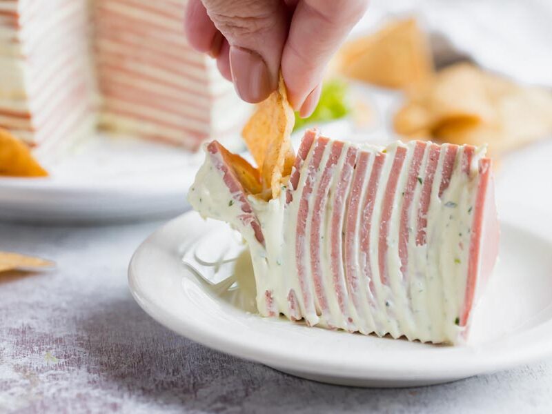 A hand dipping a chip into a slice of bologna cake.