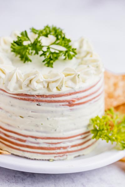 A cake made out of bologna slices and cream cheese on a plate with sprigs of parsley around.