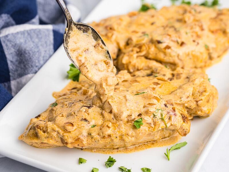 A spoon pouring creamy cheesy onion sauce over the top of cooked chicken breasts sitting on a plate.