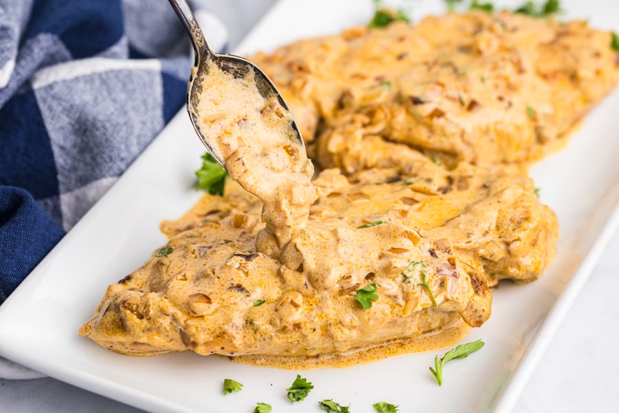 A spoon pouring creamy cheesy onion sauce over the top of cooked chicken breasts sitting on a plate.