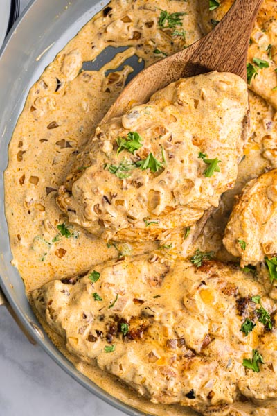 A wooden spoon holding a piece of chicken smothered in cream sauce in a skillet.