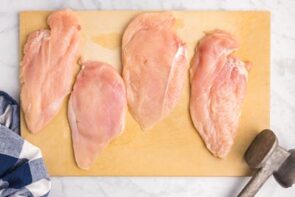 Four chicken cutlets on a sheet of parchment paper next to a meat hammer.