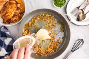 Pouring chicken broth in a skillet filled with caramelized onion and a round of boursin cheese.