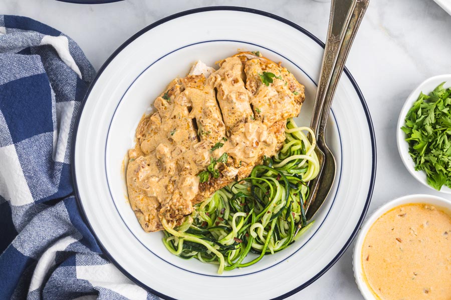 A dinner plate with serving of sliced boursin chicken covered in a creamy sauce and lying next to seasoned zucchini noodles.