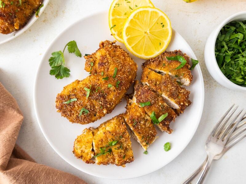 A fork tender, cut up chicken tender on a plate next to a full cutlet and some lemon slices.