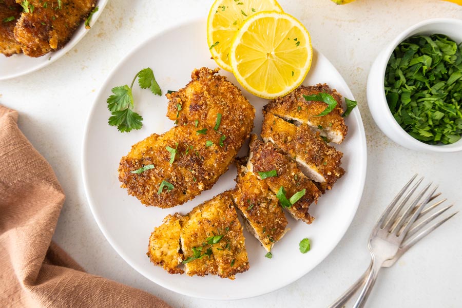 A fork tender, cut up chicken tender on a plate next to a full cutlet and some lemon slices.