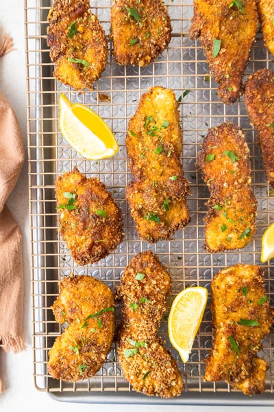 Crispy chicken cutlets on a wire rack over a sheet pan and topped with parsley and lemon wedges.
