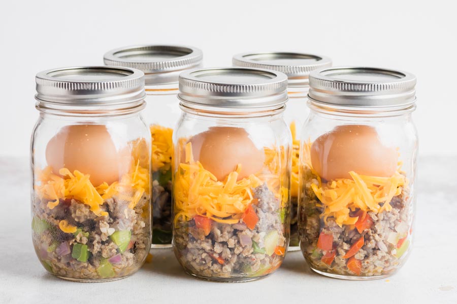 Five mason jars with layers of sausage mixture, shredded cheese and a whole egg on top.