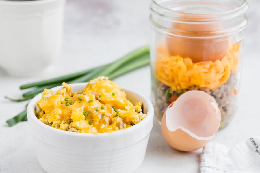 A small bowl with sausage egg scramble next to a meal prep bowl and a cracked egg shell.