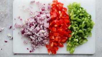 A cutting board with purple onion, red bell pepper and green bell pepper all diced in a row.