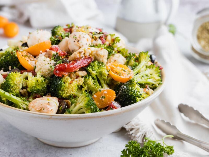 A bowl of raw broccoli salad with cauliflower, tomatoes and red pepper next to tongs and a napkin.