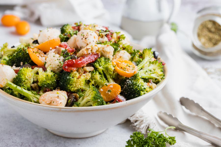 A bowl of raw broccoli salad with cauliflower, tomatoes and red pepper next to tongs and a napkin.