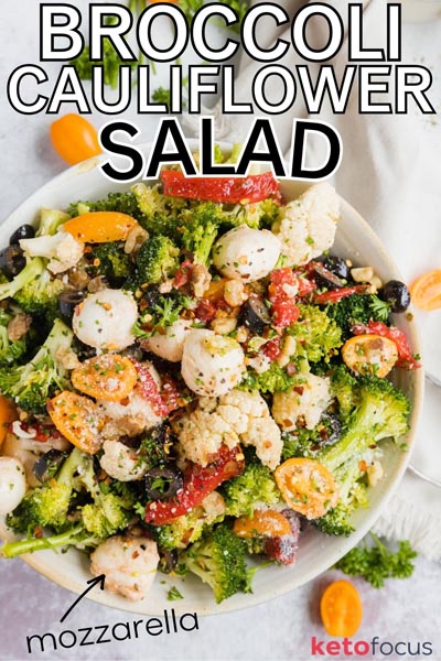 Looking into a large bowl with a broccoli cauliflower salad filled with sliced tomato, diced roasted peppers, mozzarella cheese balls and olives.