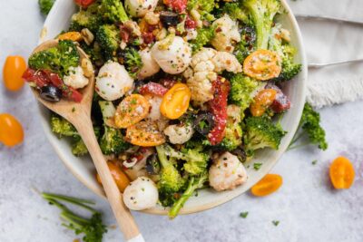 A wood spoon rests on a bowl with a colorful broccoli cauliflower dish. Broccoli, cauliflower, and olive slice and a strip of roasted red bell pepper sit in the spoon.