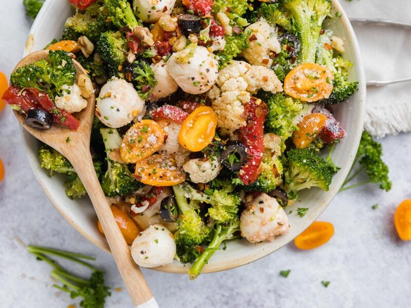 A wood spoon rests on a bowl with a colorful broccoli cauliflower dish. Broccoli, cauliflower, and olive slice and a strip of roasted red bell pepper sit in the spoon.