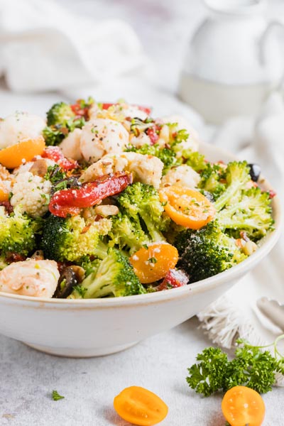 Florets of broccoli and cauliflower in a bowl with strips of red bell pepper, mozzarella cheese and golden grape tomatoes on a table.