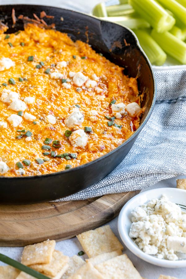 a cast iron skillet on a wooden trivet with crumbled blue cheese, celery and crackers around it. Buffalo dip sits in the skillet