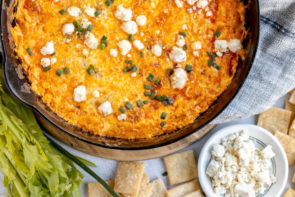 cheesy buffalo dip in cast iron skillet topped with blue cheese and chives next to a bowl of crumbled blue cheese and crackers spread around