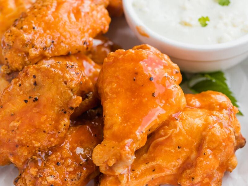 Close up of buffalo wings covered in hot sauce next to a small white bowl of blue cheese dressing.