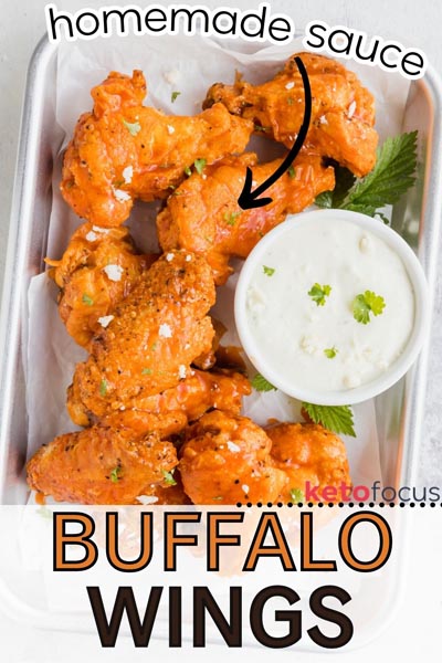 A parchment lined tray with buffalo wings topped with parsley and blue cheese crumbles next to a bowl of dressing.
