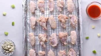 Party wings evenly spaced on a wire rack on top of a baking tray with foil. Crumbled blue cheese and hot sauce are nearby.