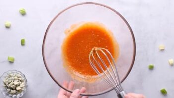 Whisking buffalo sauce in a glass bowl with chopped green onions near by.