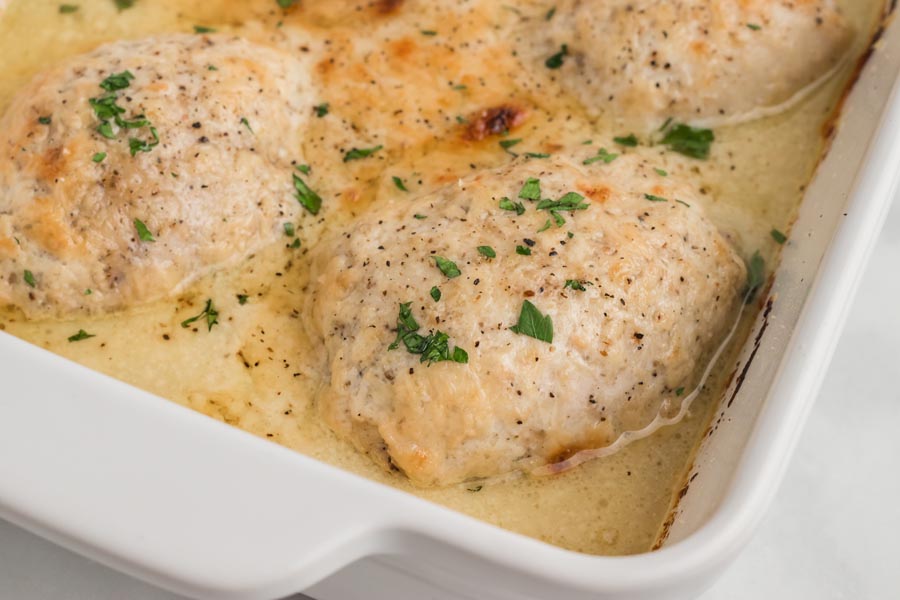 A casserole dish filled with baked chicken thighs swimming in a creamy caesar sauce and topped with black pepper and parsley.