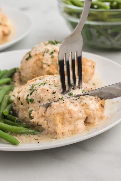 A fork and knife cutting into a baked chicken thigh next to a pile of green beans.