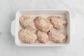 A white casserole dish with raw chicken thighs coated with shredded parmesan.