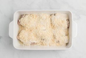 Sauce and shredded cheese all over chicken thighs sitting in a white casserole dish.