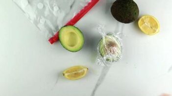 one half avocado covered in plastic wrap next to another half in front of a baggie