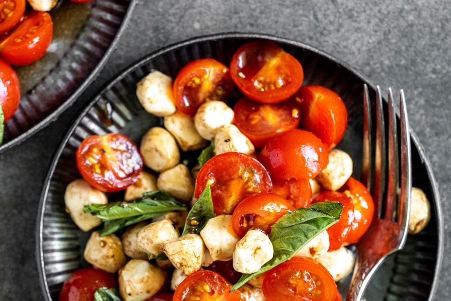 a black plate of tomatoes, mozzarella balls and basil leaves next to a fork
