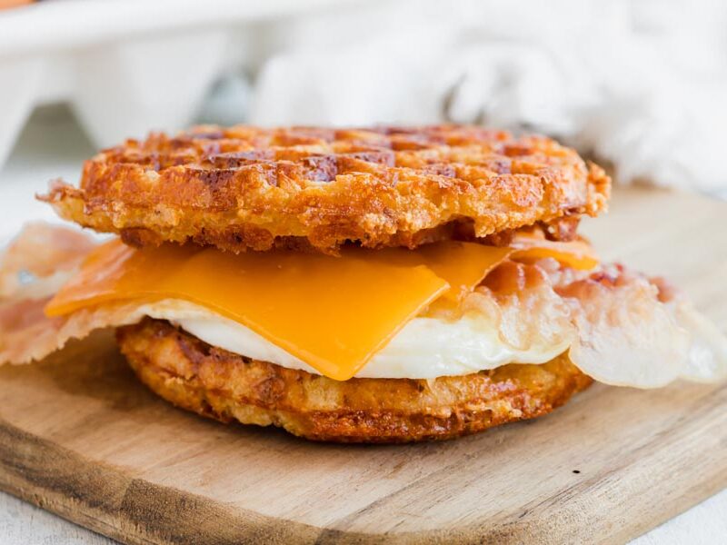A waffle sandwich with fried egg, bacon and cheddar cheese on a cutting board.