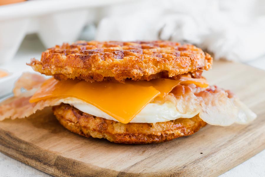 A waffle sandwich with fried egg, bacon and cheddar cheese on a cutting board.