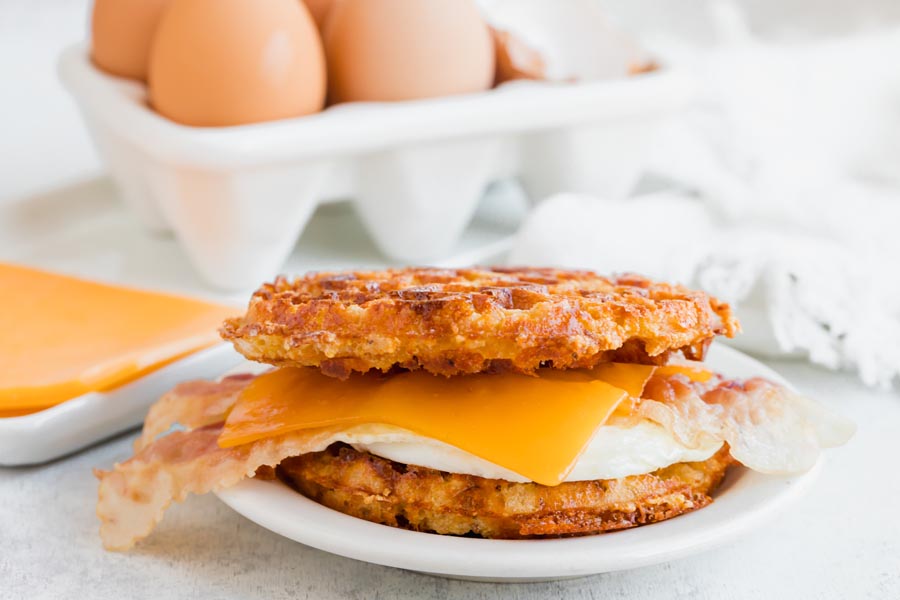 A sandwich made out of waffle, egg, cheese and bacon on a plate in front of eggs and cheese slices.
