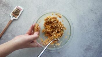 A clear mixing bowl with chaffle ingredients inside. A hand holds the bowl and a spatula sits nearby.