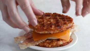 Hands placing the top waffle on a waffle sandwich with bacon, egg and cheese.