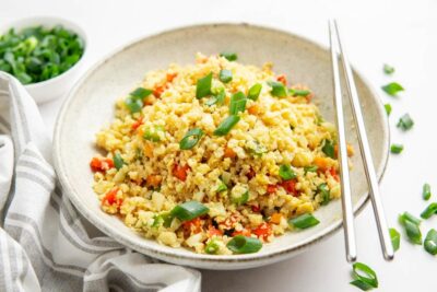 A bowl of cauliflower fried rice topped with green onions and metal chopsticks nearby.