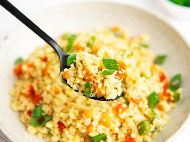 A spoon holding a bite of fluffy fried cauliflower rice mixed with vegetables.