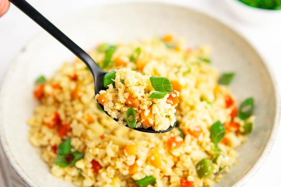 A spoon holding a bite of fluffy fried cauliflower rice mixed with vegetables.