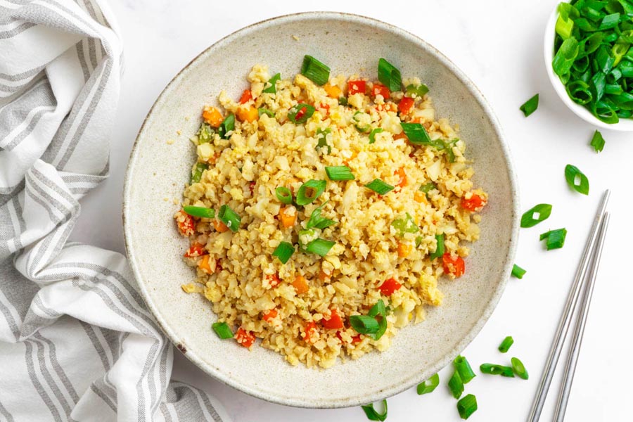 A plate of fluffy keto fried rice made with riced cauliflower, carrot, bell pepper and green onions. Green onion slices are scattered around near the plate.