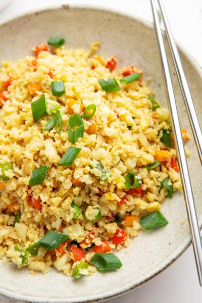 A plate of cauliflower fried rice mixed with red pepper, carrot and green onion. Chopsticks are laying on the plate.