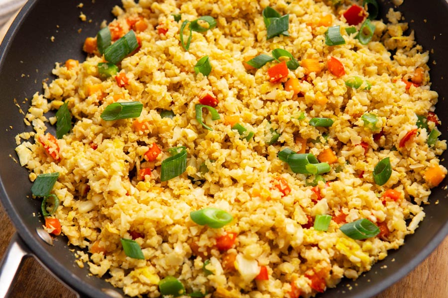 A skillet with fluffy cauliflower fried rice. The rice has chunks of carrot, red bell pepper, egg and sliced green onion.