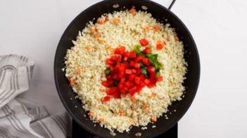 Diced red and green bell pepper sitting on top of cooking cauliflower rice in a skillet.