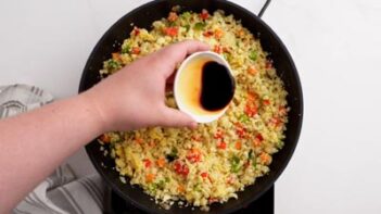 A hand pouring soy sauce into fried rice inside a skillet.