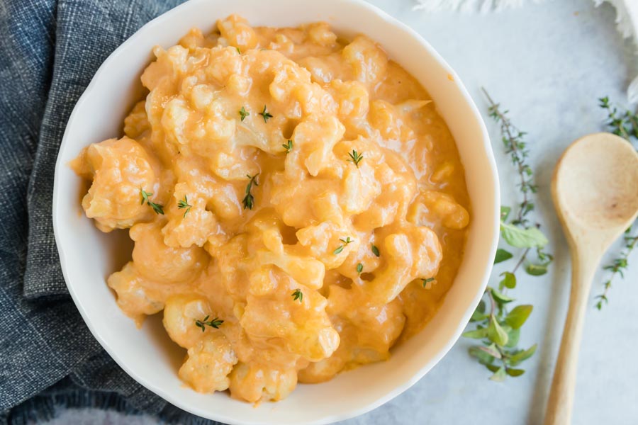 a bowl of keto cauliflower mac and cheese next to a spoon and fresh herbs