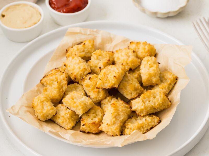 A bowl of cheesy cauliflower tots on a plate with mustard and ketchup in the background.