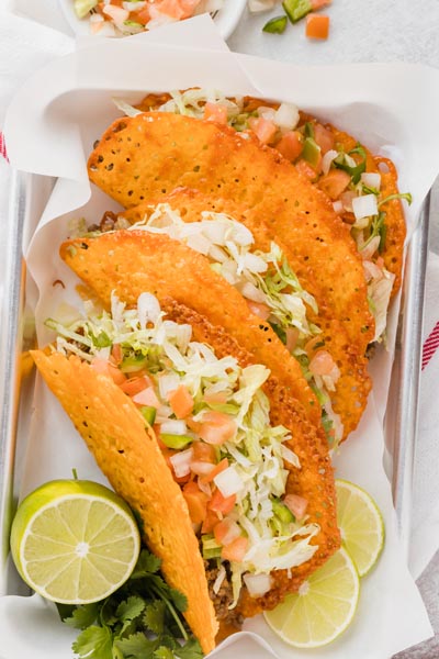 Three tacos filled with ground beef, lettuce and tomato and a taco shell made out of cheese on a parchment lined tray with limes slices nearby.